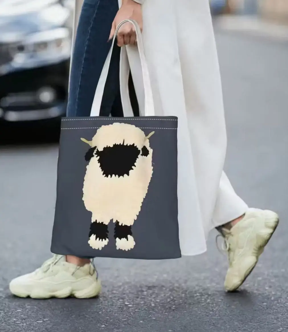 Gray tote bag with a cute sheep design held by a person wearing white pants and sneakers.